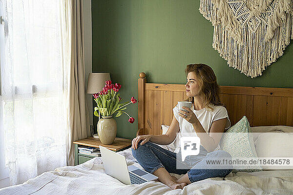 Thoughtful freelancer with coffee cup sitting on bed at home