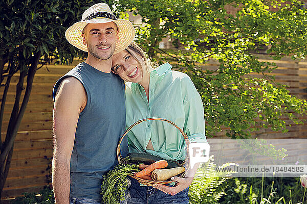 Happy young couple standing with basket of vegetables in garden