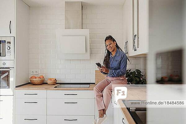 Smiling mature woman sitting on kitchen counter with smart phone at home