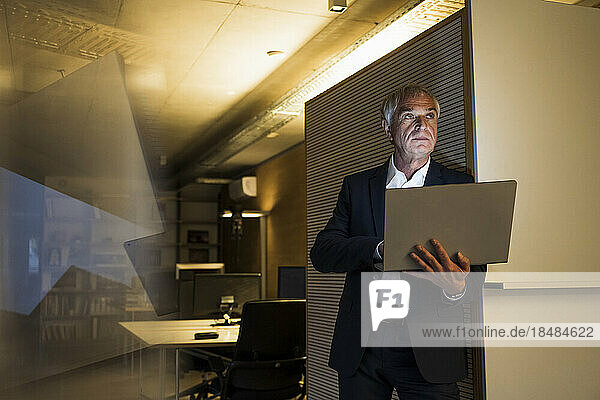 Contemplative businessman with laptop standing near wall at office
