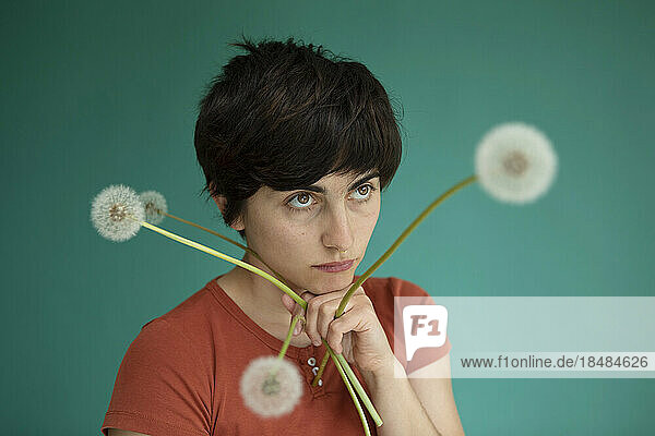 Thoughtful woman with hand on chin holding dandelions against green background