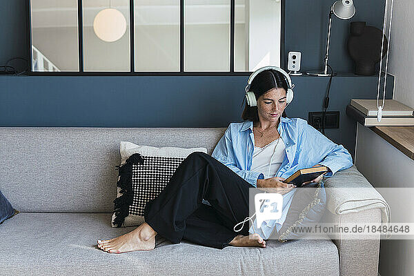Young woman with book listening to music through headphones at home