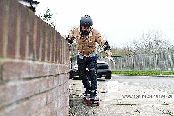 Mature man learning skateboarding taking support of wall on footpath