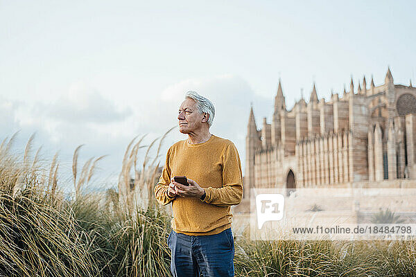Smiling senior man with smart phone standing in front of building