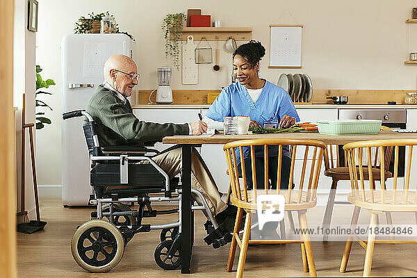 Smiling senior man sitting on wheelchair with caretaker cutting vegetables at home