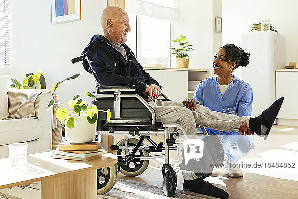 Lächelnder Physiotherapeut untersucht zu Hause das Bein eines älteren Mannes im Rollstuhl