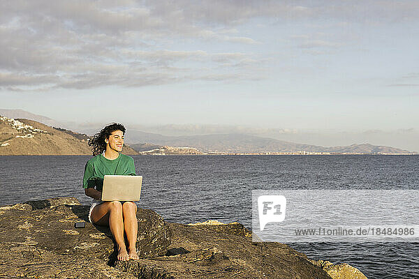 Smiling woman with laptop sitting on rock by sea