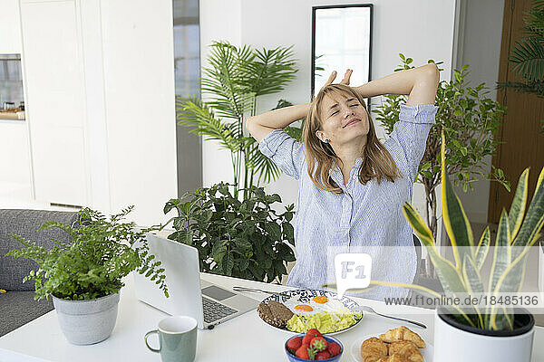 Relaxed freelancer with laptop and breakfast on table at home office