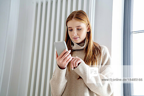 Smiling teenage girl using smart phone in front of radiator at home