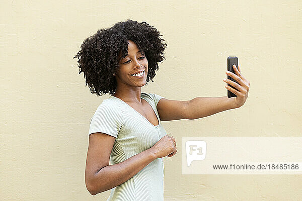 Smiling young woman taking selfie using smart phone by beige wall