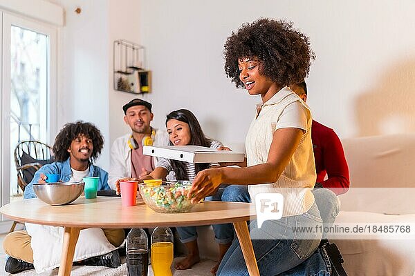 Eine Gruppe multiethnischer Freunde sitzt auf einem Sofa  isst Pizza und trinkt alkoholfreie Getränke bei einer Party zu Hause  ein junger Mann kommt mit der Pizza von der Straße