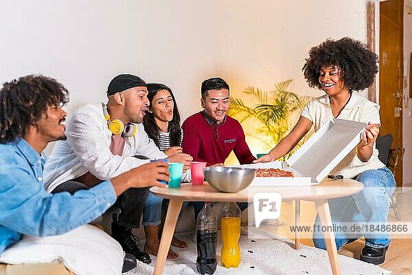 Eine Gruppe multiethnischer Freunde sitzt auf einem Sofa  isst Pizza und trinkt alkoholfreie Getränke bei einer Party zu Hause  ein junger Mann kommt mit einem Pizzakarton von der Straße