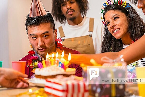 Multiethnische Gruppe von Freunden bei einer Geburtstagsfeier auf dem Sofa zu Hause mit einem Kuchen und Geschenken  Anzünden der Kerzen