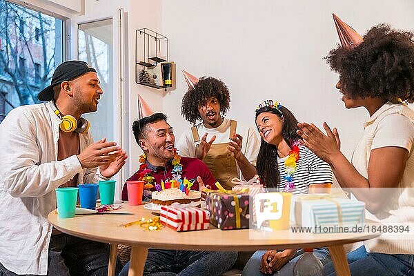 Multiethnische Gruppe von Freunden bei einer Geburtstagsfeier auf dem Sofa zu Hause mit einem Kuchen und Geschenken  die ihm ein Geburtstagsständchen singen