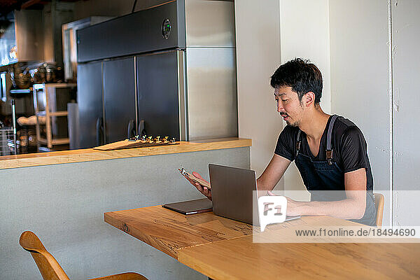 A man seated at a table using a laptop computer  owner and manager of a small restaurant.