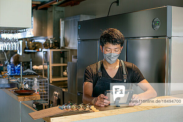 A man wearing a face mask at the counter of a restaurant kitchen  using a digital tablet  the owner or manager.