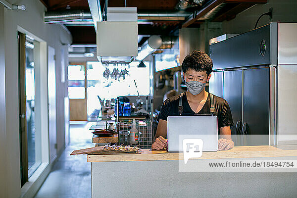 A man in a face mask in a restaurant kitchen  using a laptop  the owner or manager.