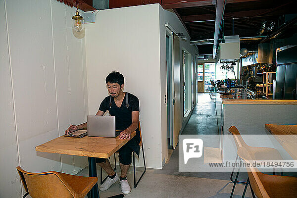 A man seated at a table using a laptop computer  owner and manager of a small restaurant.