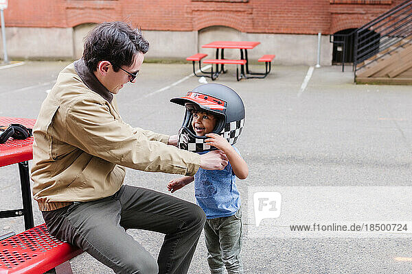 Smiling man helping toddler put motorcycle helmet on outside