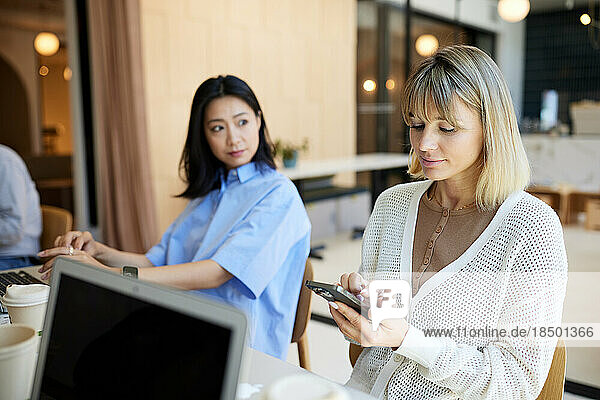 Businesswoman using smart phone sitting by female colleague in cafe