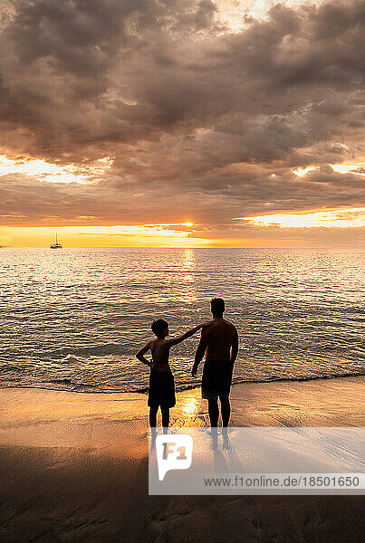 Father and son standing on a sandy beach watching the sunset together.