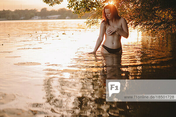 Topless woman covering her breast while standing in lake at sunset