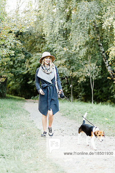 young woman in a coat and hat walks a beagle dog in the park