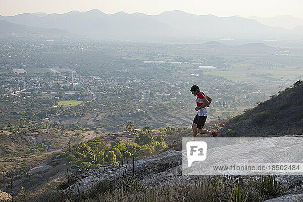Athletic male trail runner flying down rocky terrain in the arid mountains of El Arenal  Hidalgo  Mexico  as the sun sets ins the distance and lights up the town below.