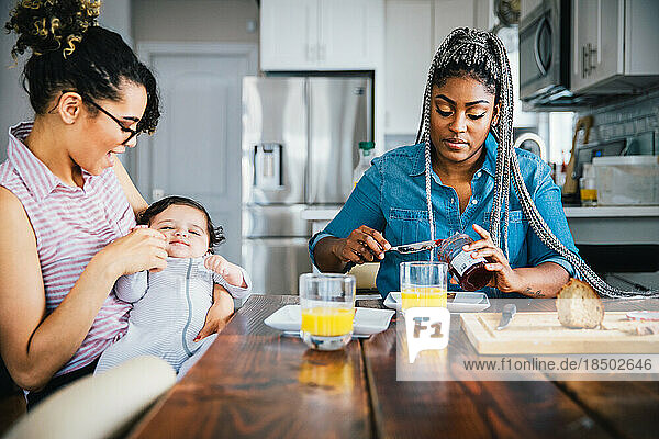 Happy mother playing with baby while girlfriend preparing breakfast at table