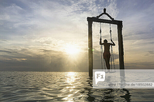 Rear view of young man standing on rope swing at beach against sunset  Gili Trawangan  Lombok  Indonesia