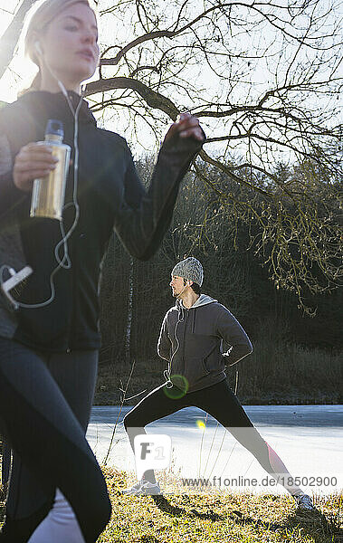 Woman jogging while man is stretching on fitness trail in front of frozen lake