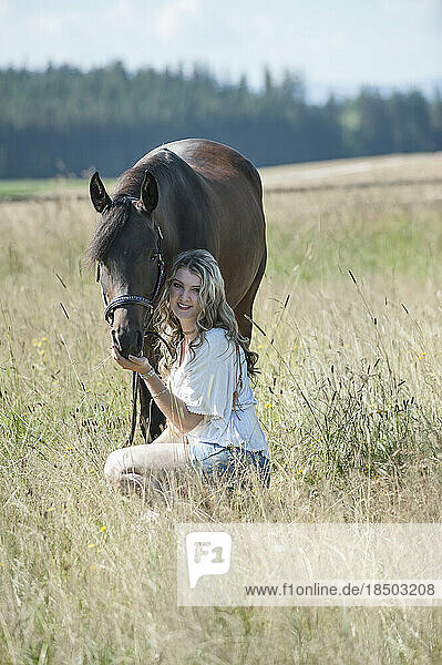 Young woman stroking her brown horse sitting on meadow and smiling  Bavaria  Germany
