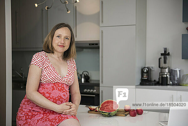 Portrait of a pregnant woman sitting on table in the kitchen and smiling  Munich  Bavaria  Germany