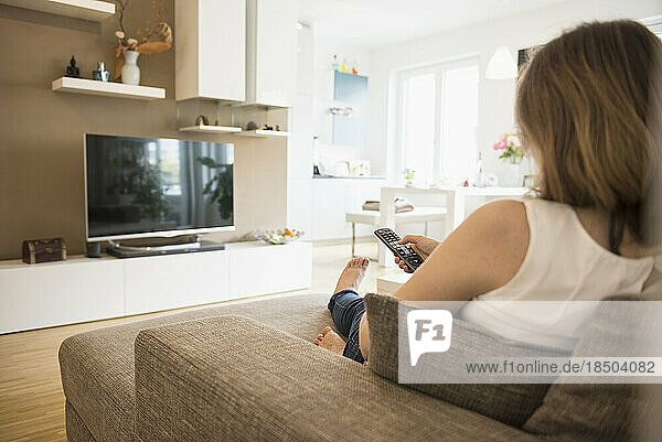 Pregnant woman sitting on sofa and watching TV with remote control  Munich  Bavaria  Germany