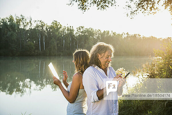 Couple at a river using smart phone and digital tablet  back to back