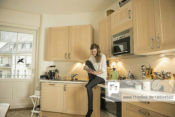 Young woman using digital tablet and sitting on kitchen worktop  Munich  Bavaria  Germany