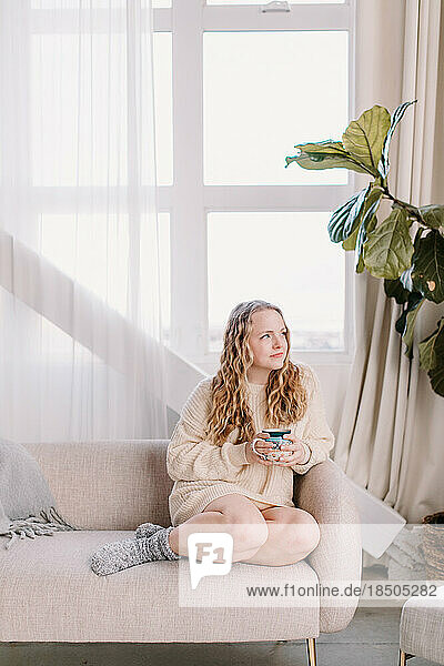 Thoughtful young woman in cozy sweater on couch looking out window