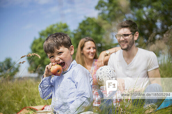 Little boy eating an apple on meadow in the countryside with family in the background  Bavaria  Germany