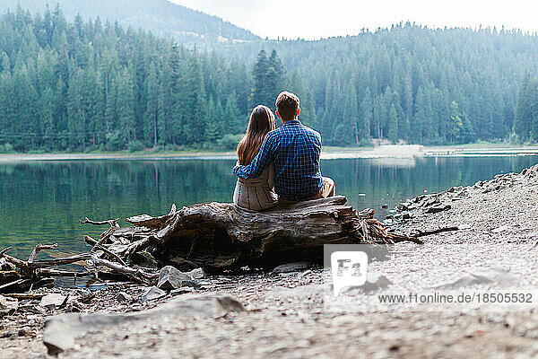 man and a woman are sitting hugging near a forest lake