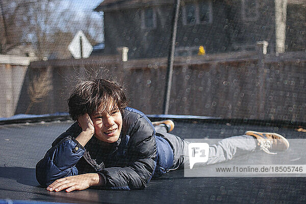 A smiling boy lays on a trampoline outside in winter