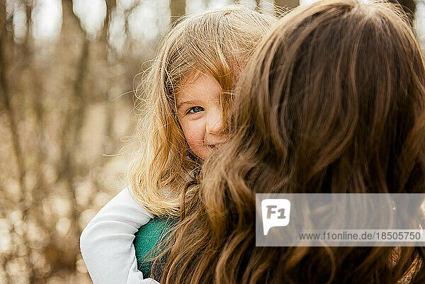 Smiling girl peeks over mothers shoulder outside in Fall woods