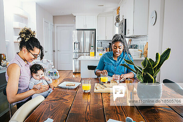 Happy mother playing with baby boy while girlfriend preparing breakfast at table