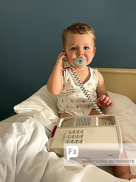 baby boy with pacifier and telephone on bed looking in to camera