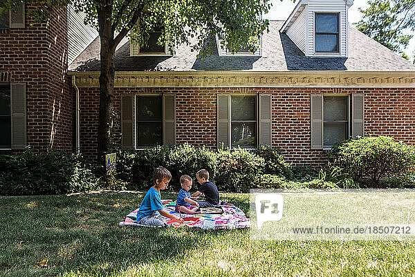 3 boys on a quilt in front yard of brick house doing school work