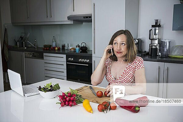 Pregnant woman talking on smartphone and sitting on chair with fresh vegetables in the kitchen  Munich  Bavaria  Germany