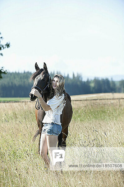 Young woman stroking her brown horse standing on meadow and smiling  Bavaria  Germany