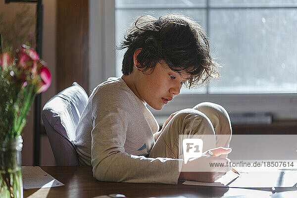 A boy with tousled hair sits at table writing on paper with pencil