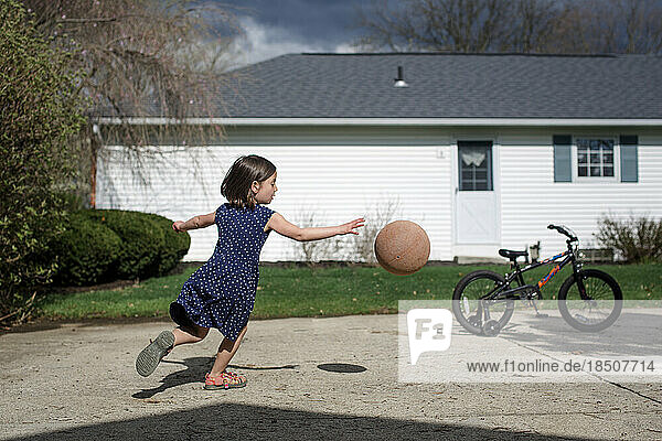 A smal girl chases a basketball in driveway against cloudy sky
