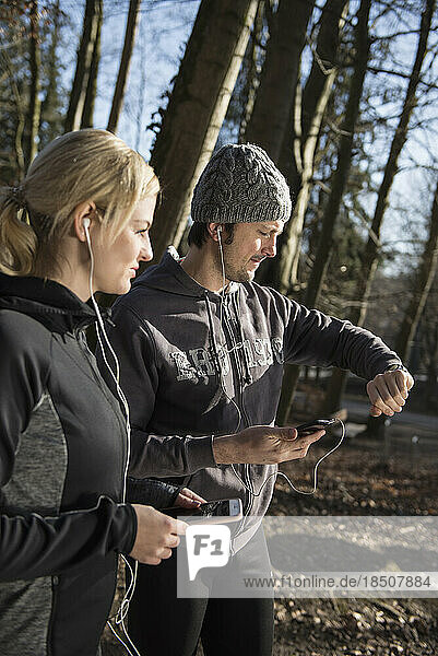 Man and woman in sportswear holding smart phone with headphones and checking the time in forest