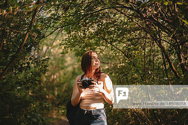 Female traveler with camera looking away while standing in forest